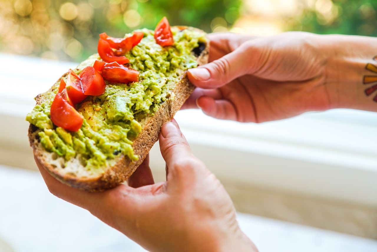 about-us-02 Close-up of avocado toast topped with cherry tomatoes being held by tattooed hands.