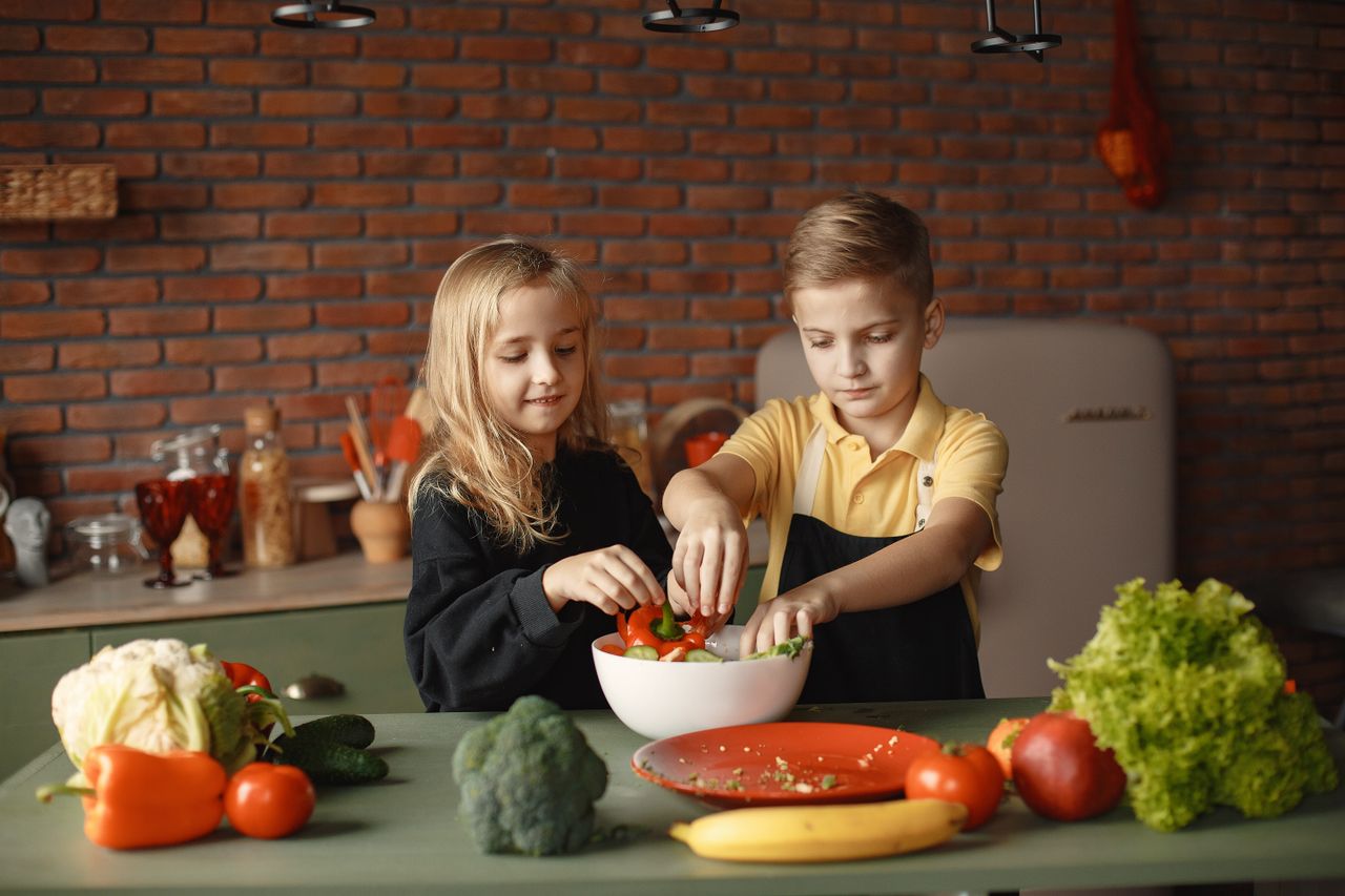 about-us-02 Cheerful girl helping little boy in apron preparing salad while standing in kitchen near table with mix of fresh vegetables behind brick wall